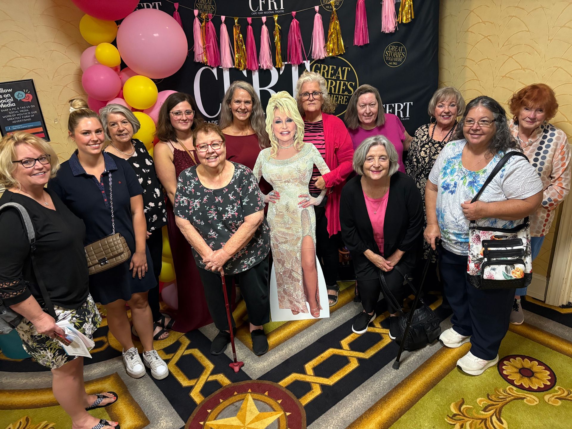 Group of women pose with a Dolly Parton cutout, balloons, and event banner.