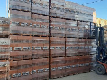 Ladrillos apilados en palés en un almacén, con una carretilla elevadora a la derecha.