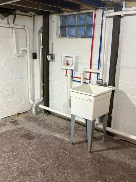 Basement utility sink beside exposed pipes, cinderblock walls, and a small window.