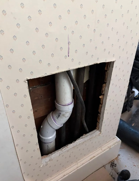 Open wall panel showing white plumbing pipes behind patterned wallpaper