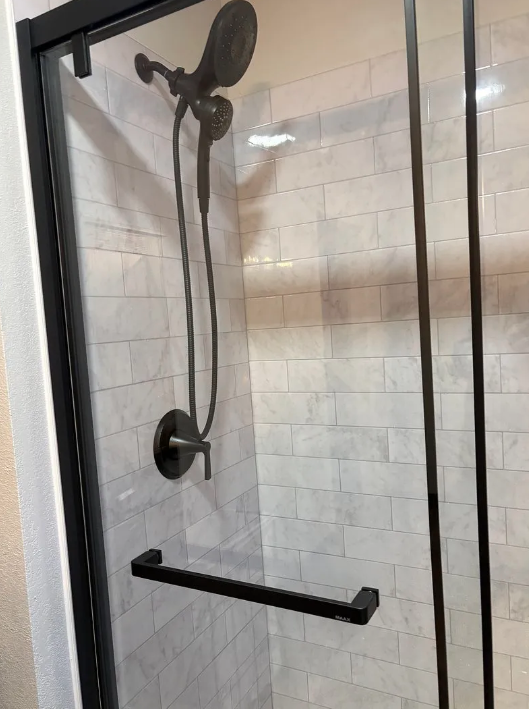Black shower with glass door, wall-mounted showerhead, and horizontal grab bar in a tiled bathroom.