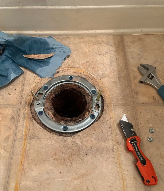 Bathroom floor with exposed toilet flange, tools, and cleaning cloths around it