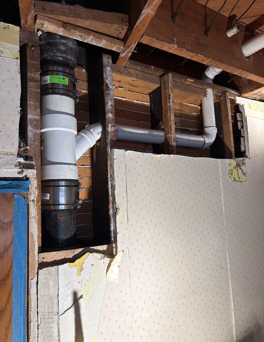 Exposed white PVC pipes and ducting inside a wall cavity above beige drywall, with unfinished wood framing.