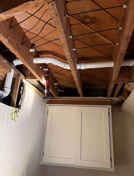 Damaged ceiling with exposed wooden rafters above a white cabinet in a room