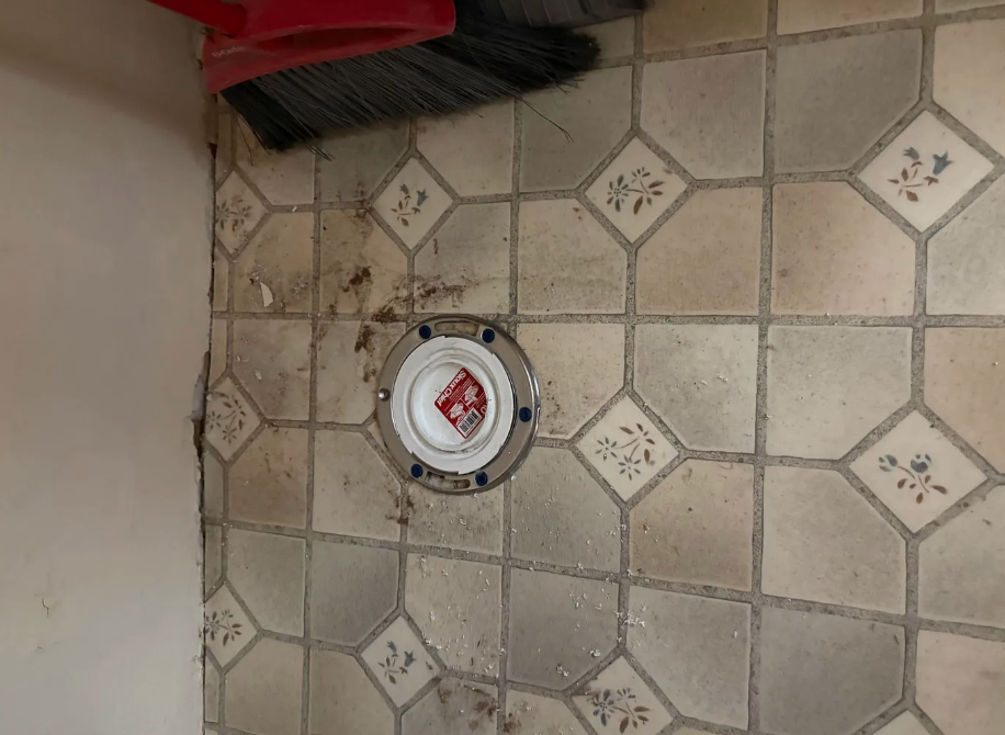 Tile floor with a round floor drain under a red brush or broom handle in the corner