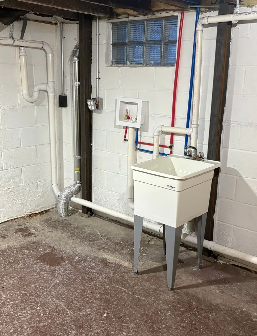 Basement utility sink beneath a small window, with exposed pipes and white-painted block walls.