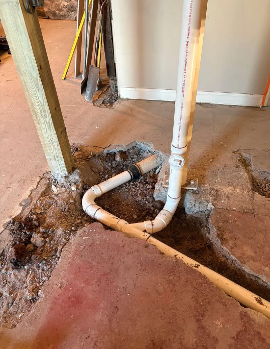 Exposed white plumbing pipes and a floor drain in a partially demolished room with dirt and debris