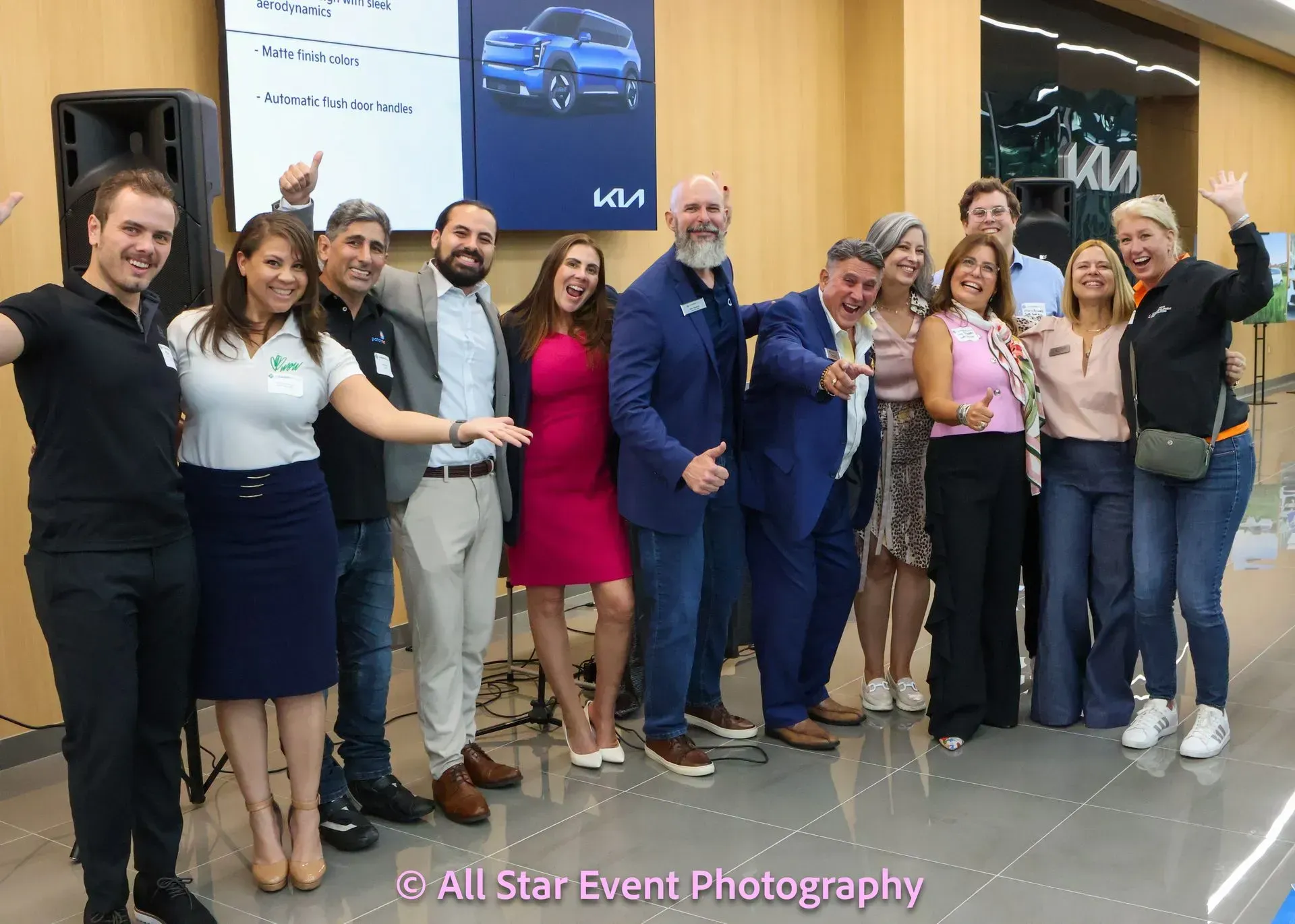 Group of people smiling and waving in a Kia showroom; a car ad is visible.