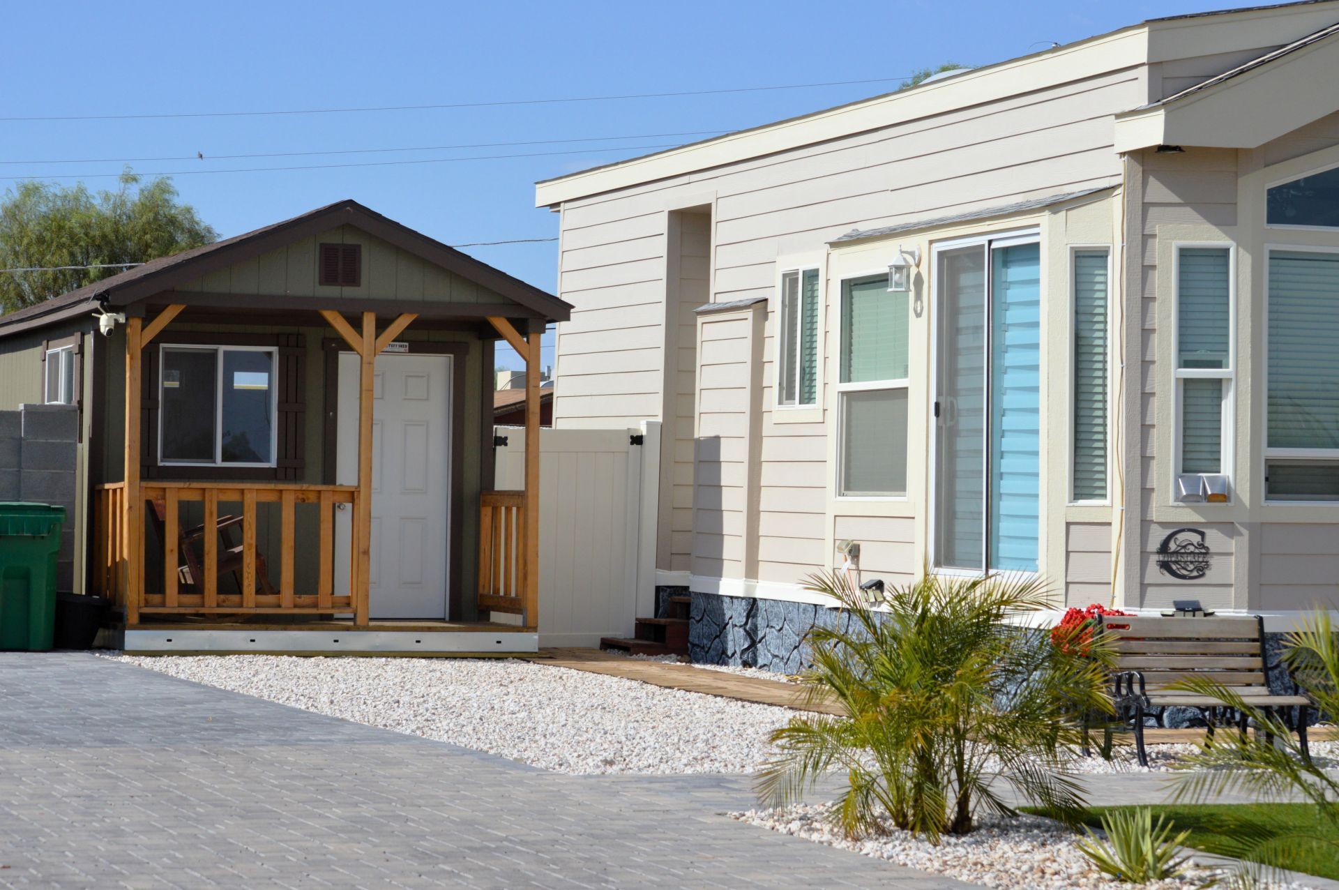 Tiny cabin with wooden porch next to a light-colored building with sliding doors; parked on white gravel.