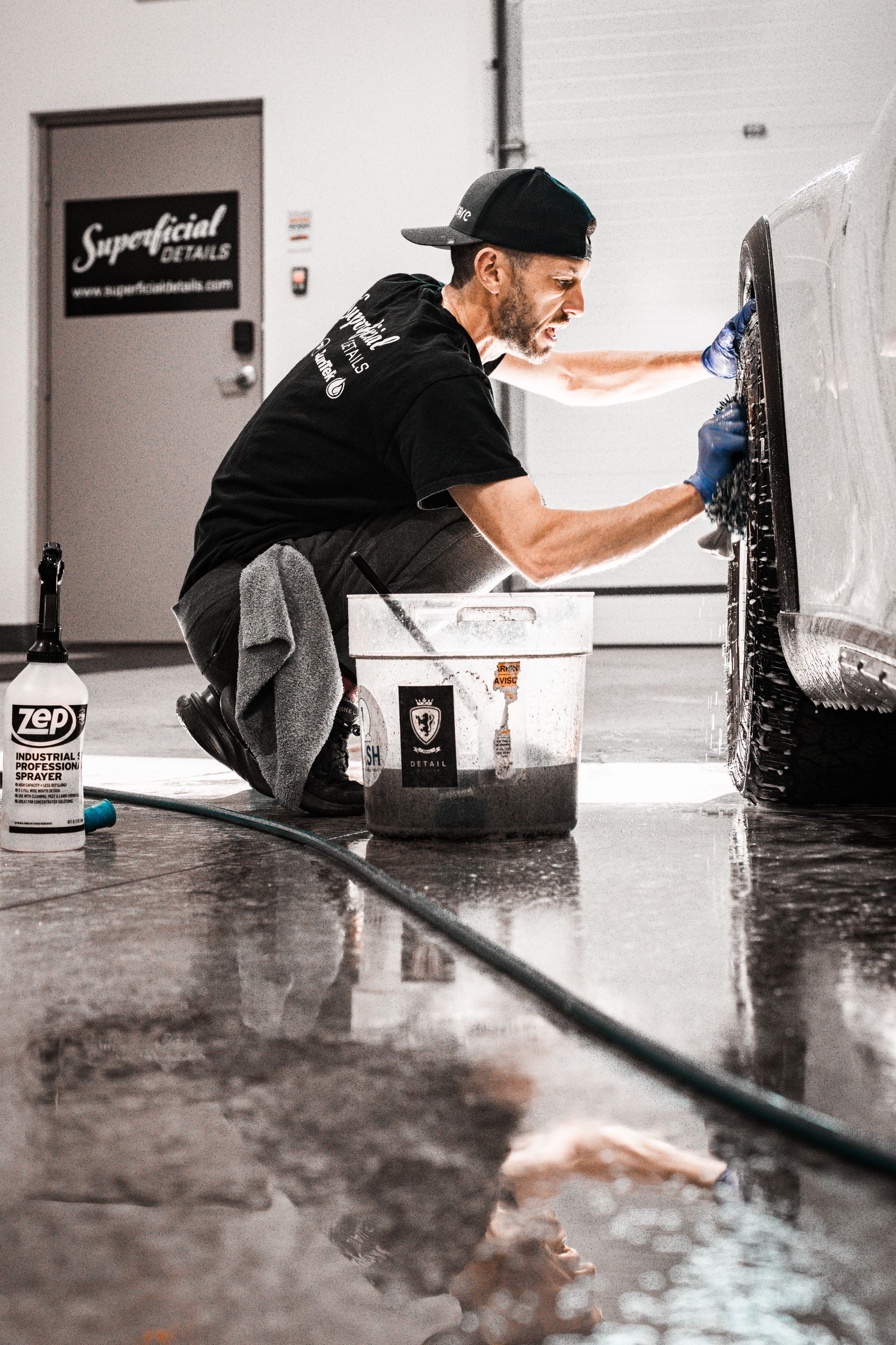 Person kneeling and scrubbing a black car in a garage, with cleaning buckets and a hose nearby