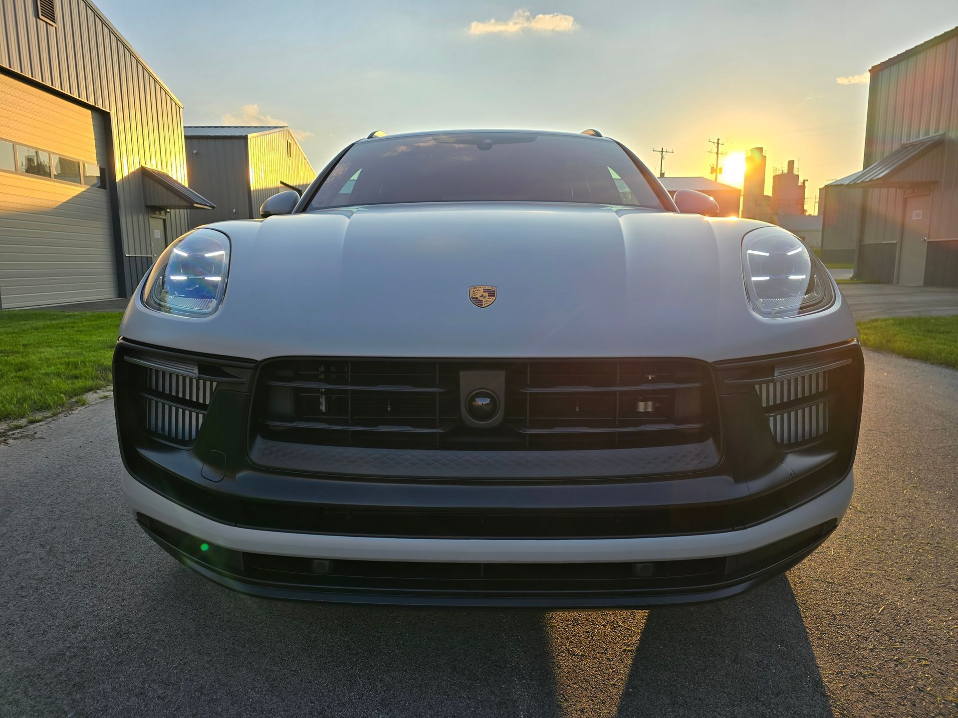 Front view of a silver Porsche parked on a street at sunset, with warehouses on both sides.