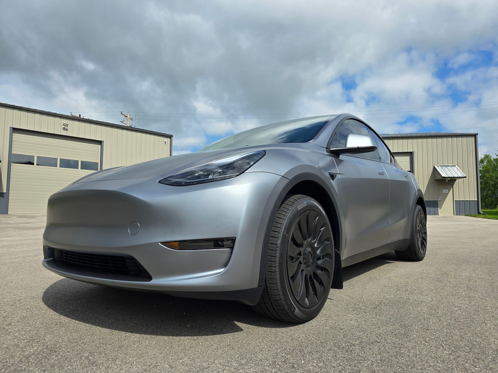 Silver Tesla Model Y parked outside industrial buildings under a cloudy sky.