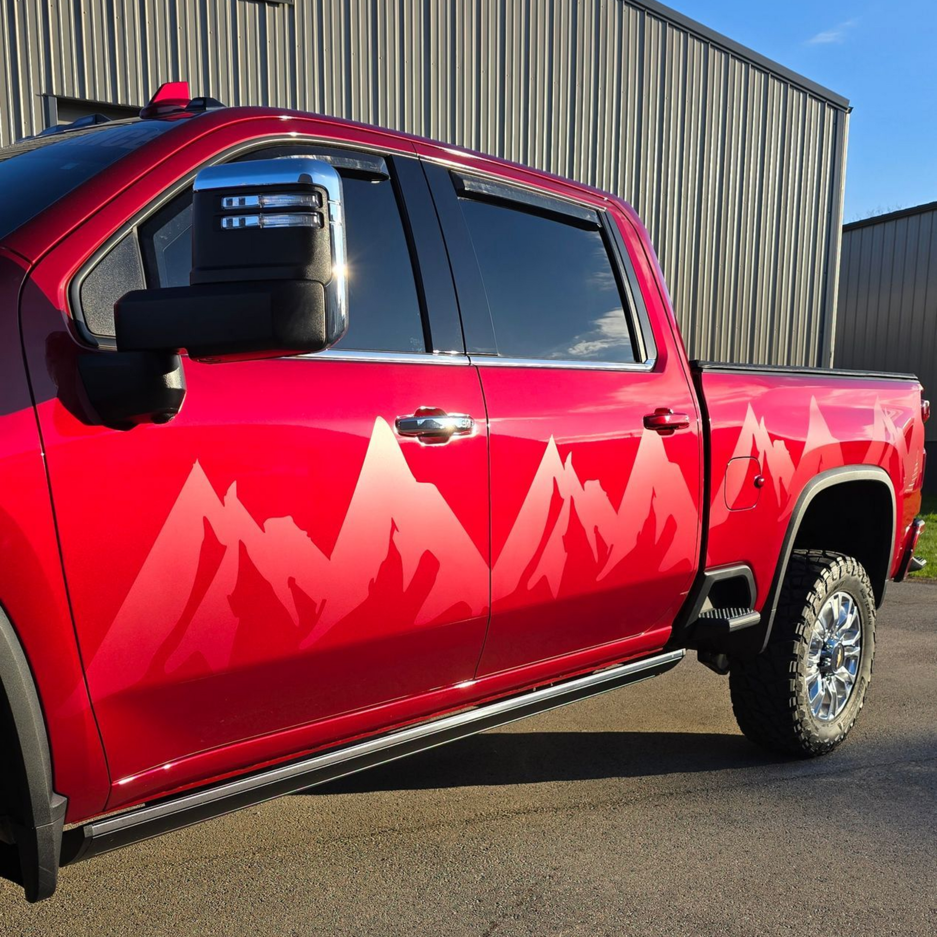 Red pickup truck with red mountain graphics, parked beside a building in sunlight