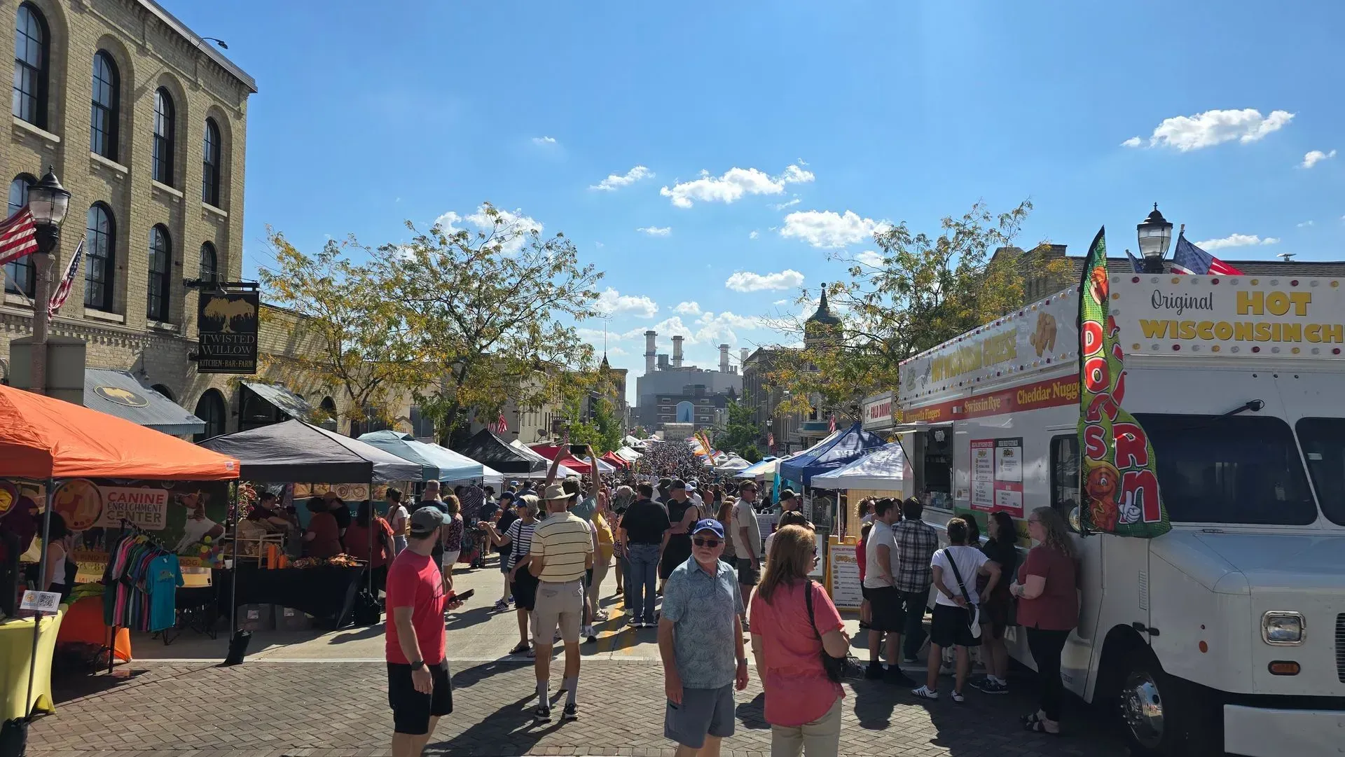 Busy outdoor market street with stalls, pedestrians, and a white food truck on a sunny day