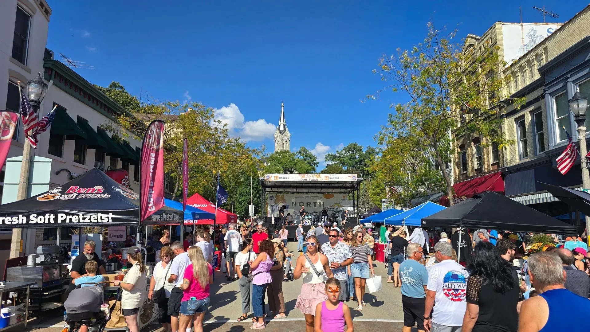 Crowded outdoor market street with colorful tents, people walking, and a clear blue sky