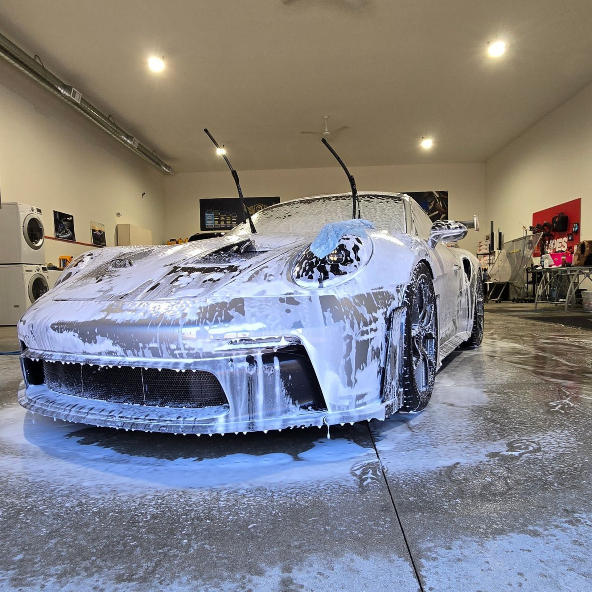 Person washing a black car with a blue sponge, covered in soap suds and water splashes