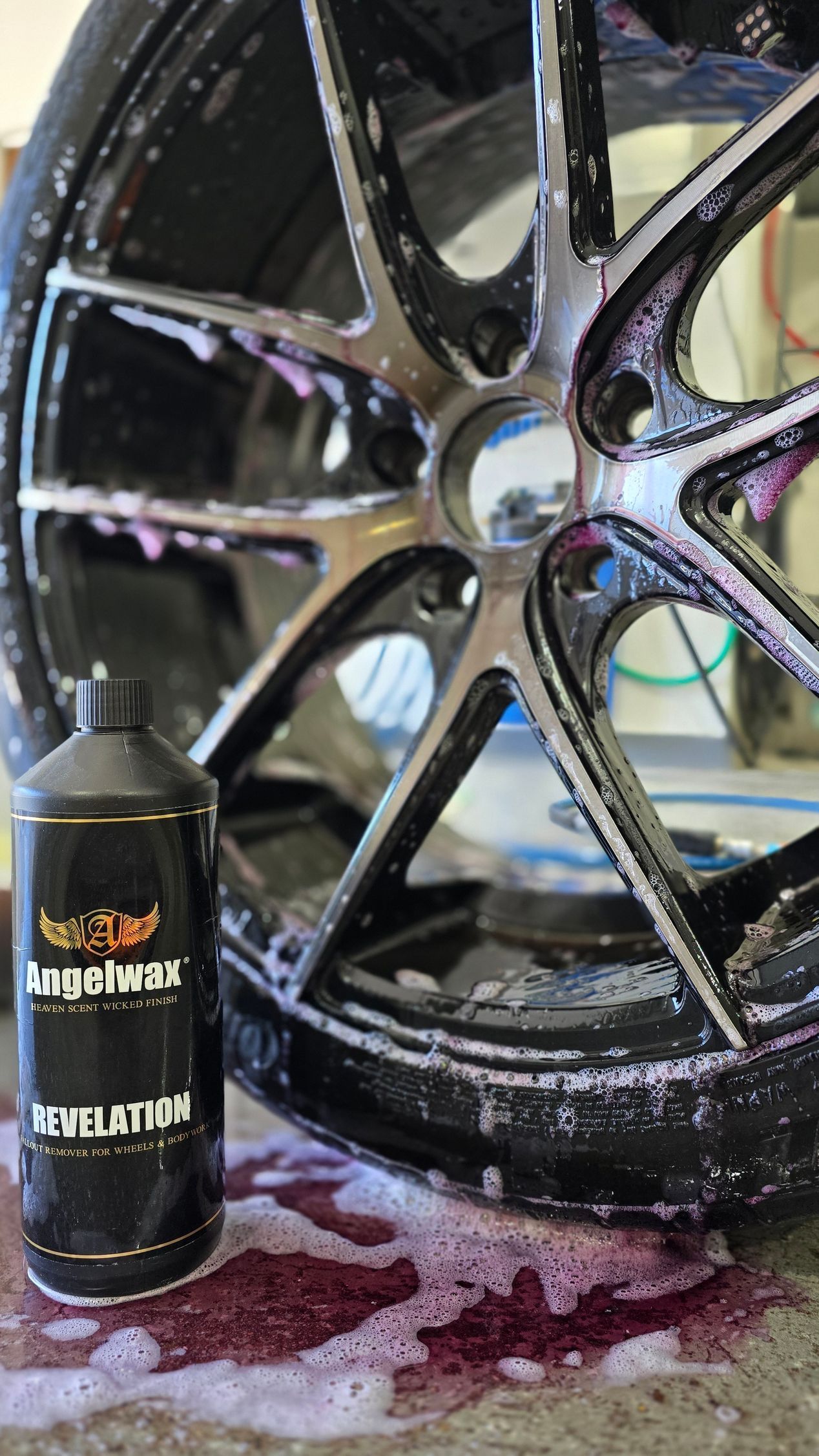 Black alloy wheel being cleaned with a detail spray bottle, with soapy foam on the ground.