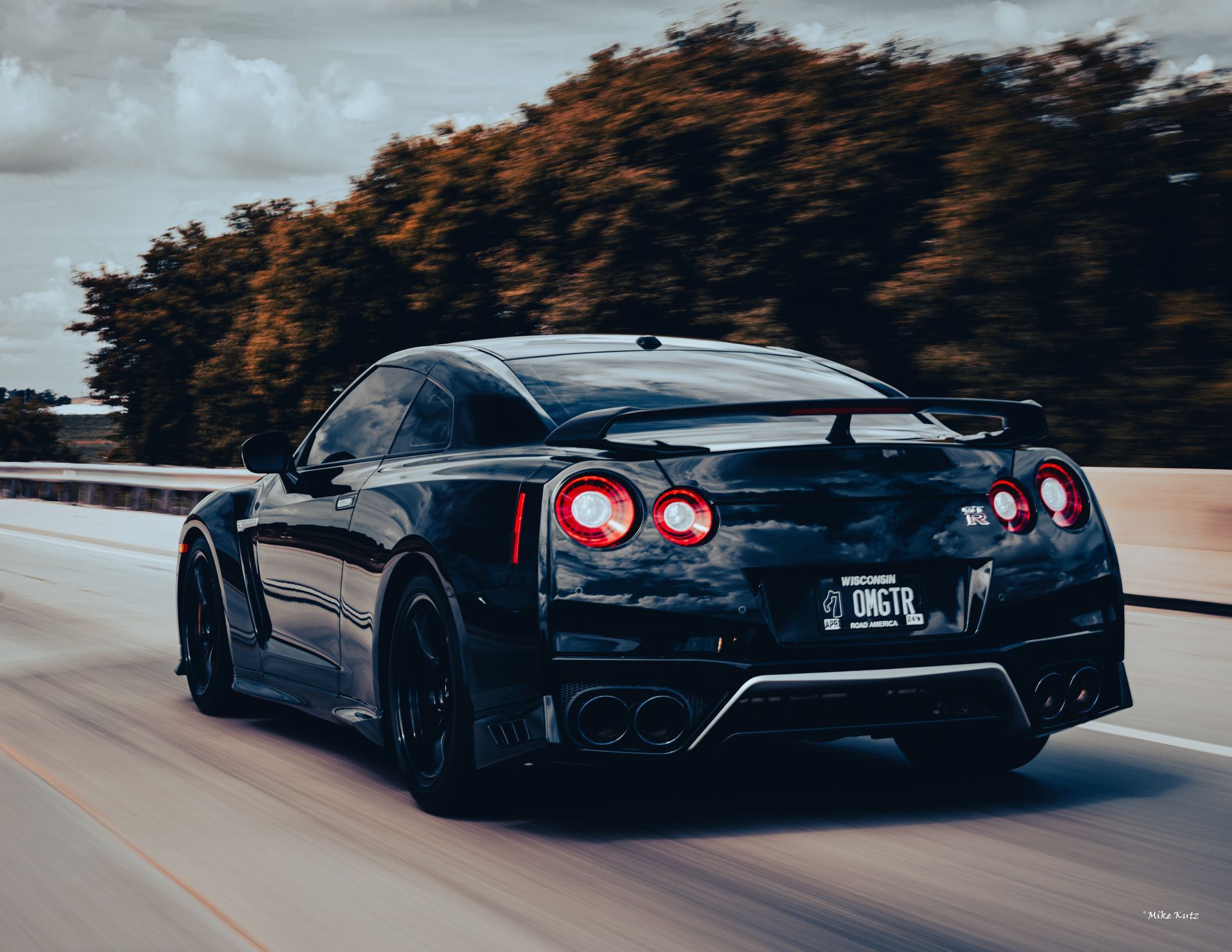 Black sports car driving on a road, viewed from the rear with taillights glowing and trees blurred in the background.