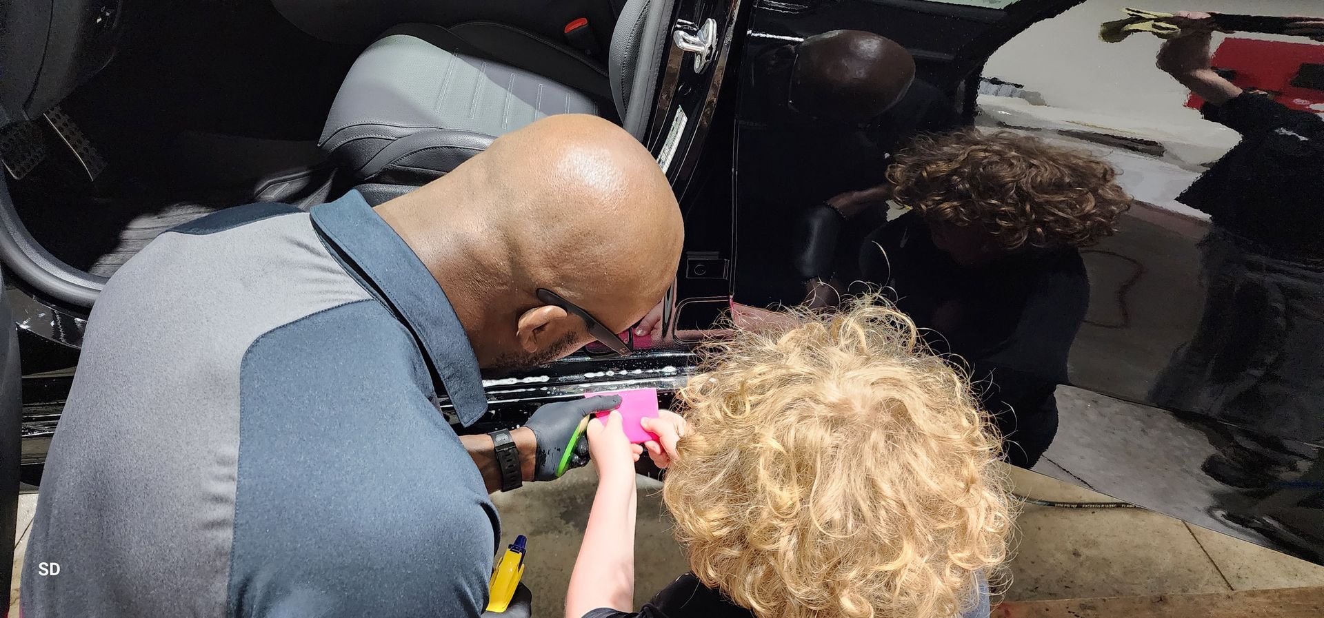Two people lean over an open car trunk, inspecting or working on colorful items inside.