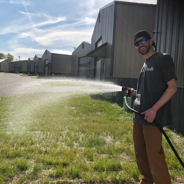 Man watering grass outside gray industrial buildings on a sunny day