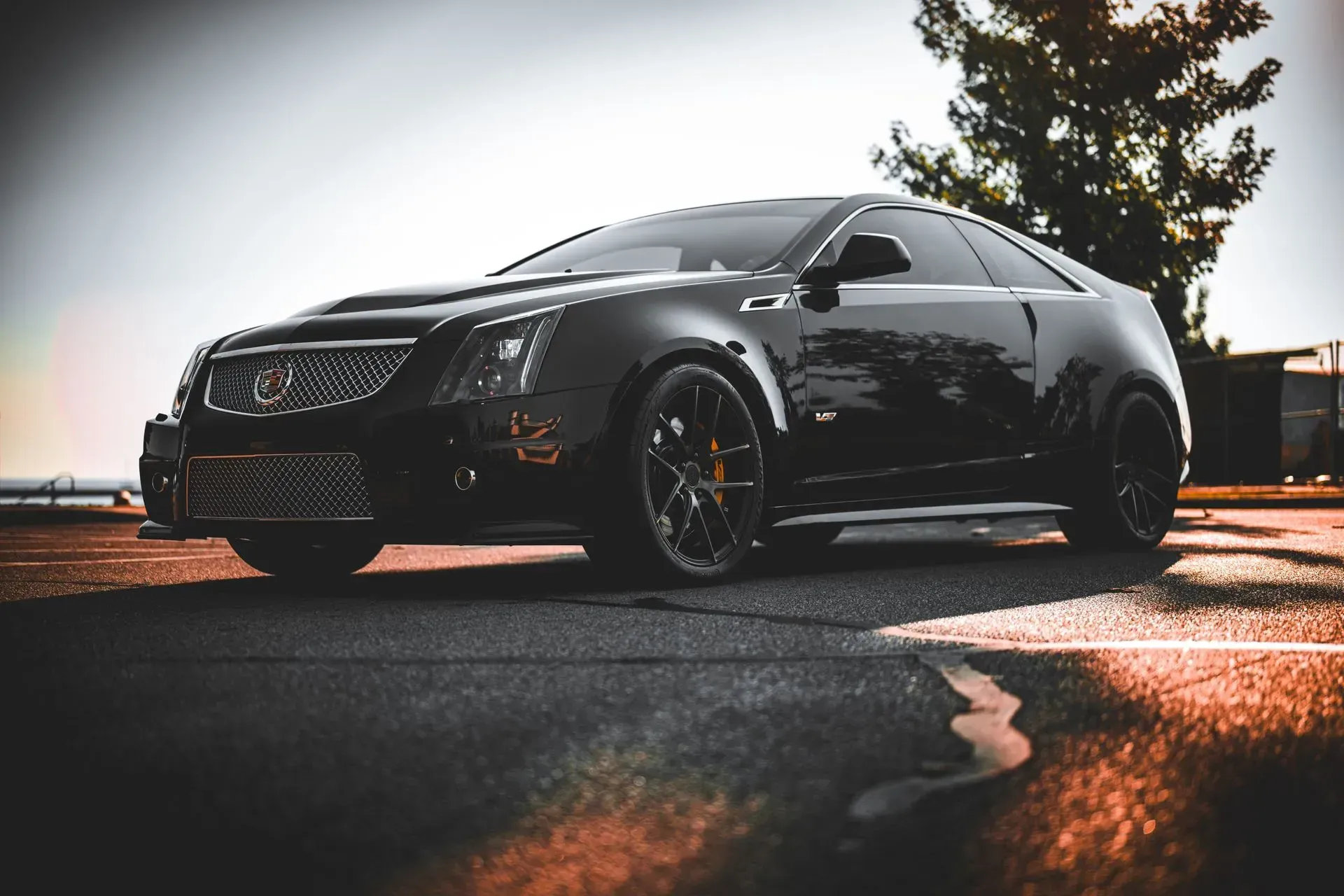 Black sports sedan parked on a wet road at sunset, with trees silhouetted in the background.