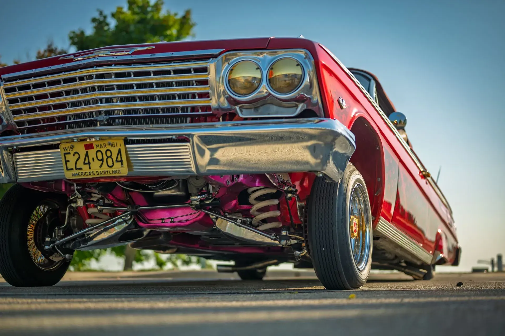 Red lowrider classic car with chrome grille and pink undercarriage, viewed from a low angle on pavement.