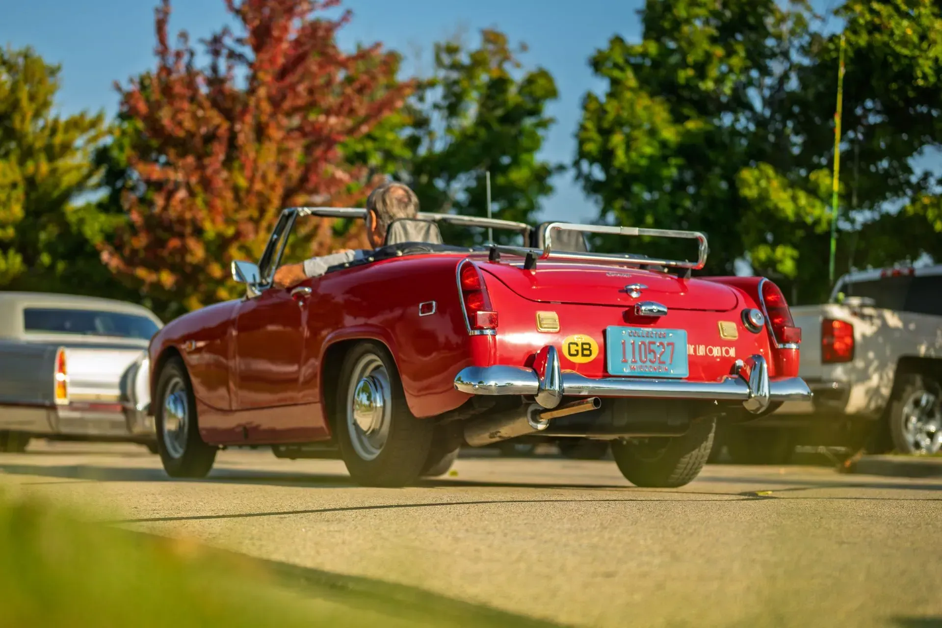 Red convertible driving on a sunlit road with autumn trees and parked cars nearby