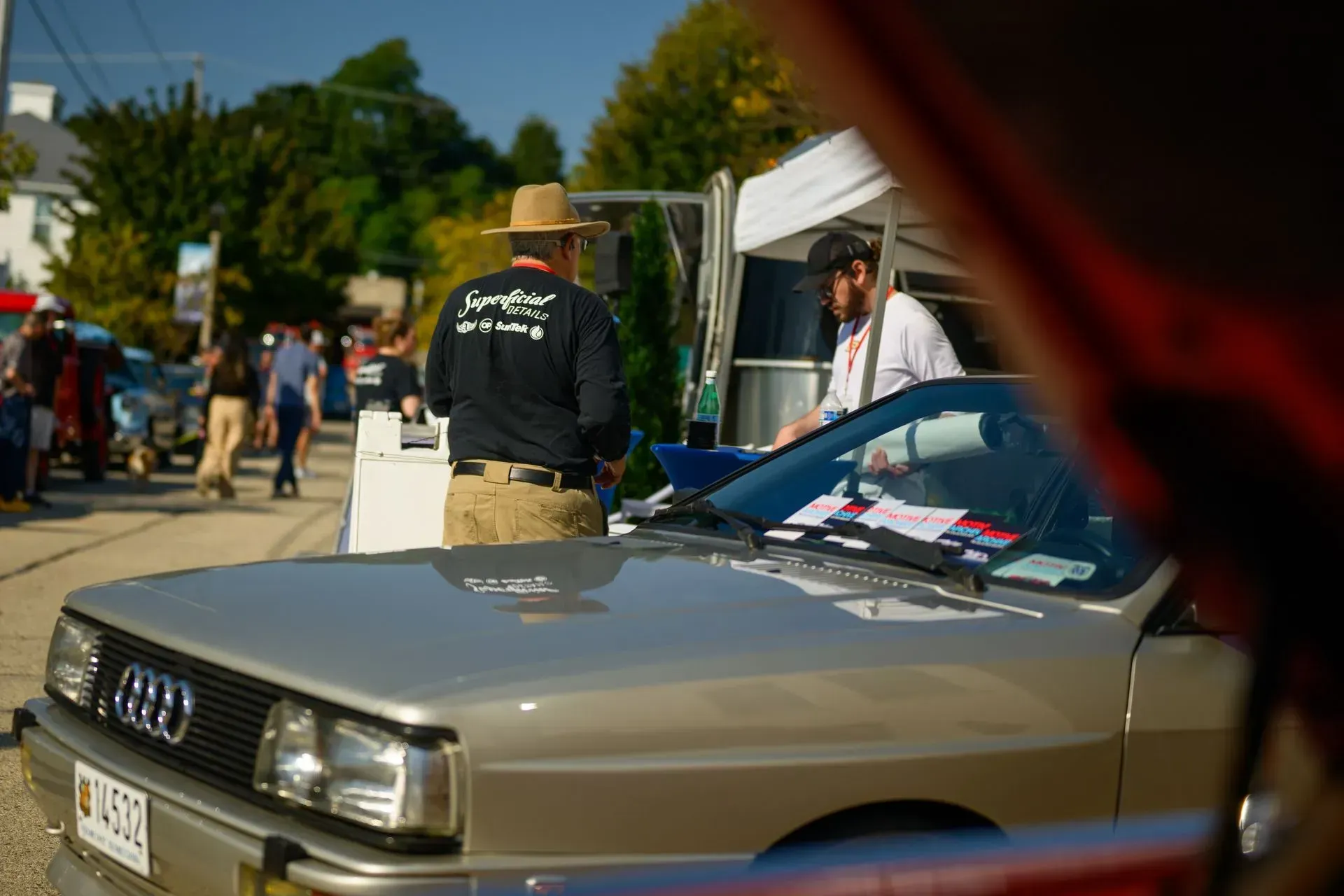 Classic beige car at an outdoor event, with people and tents in the background