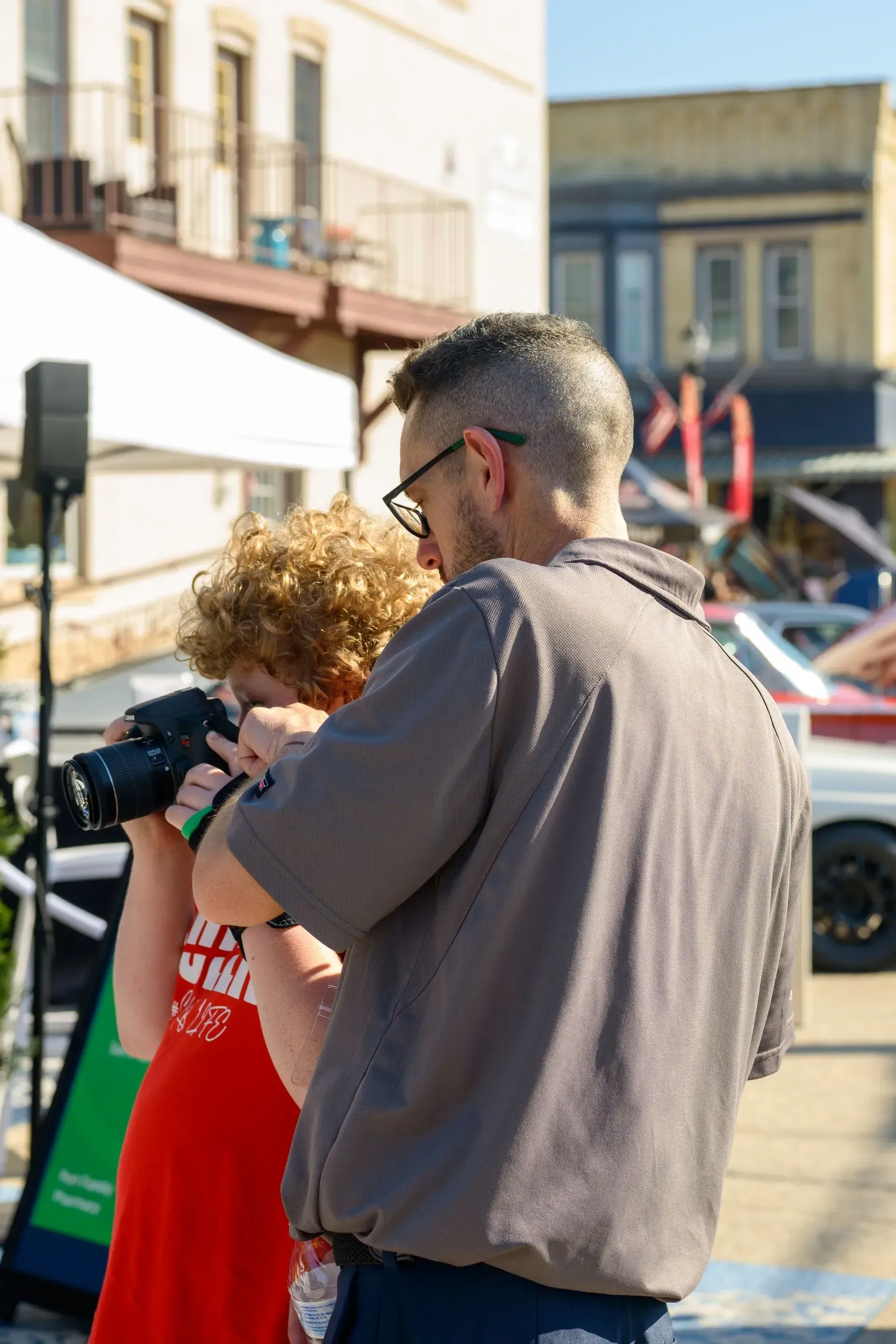 Two people standing close together outdoors, one holding a camera in a sunny street scene