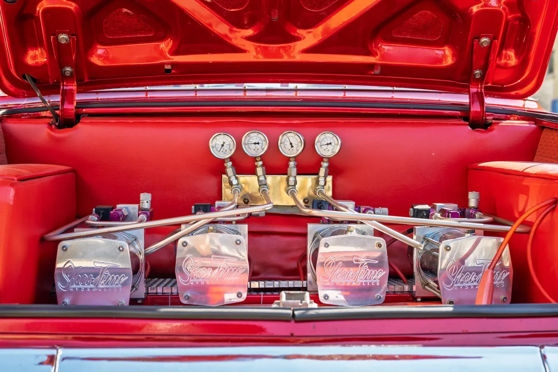 Red engine bay with polished chrome components, four gauges, and exposed fuel lines under an open hood