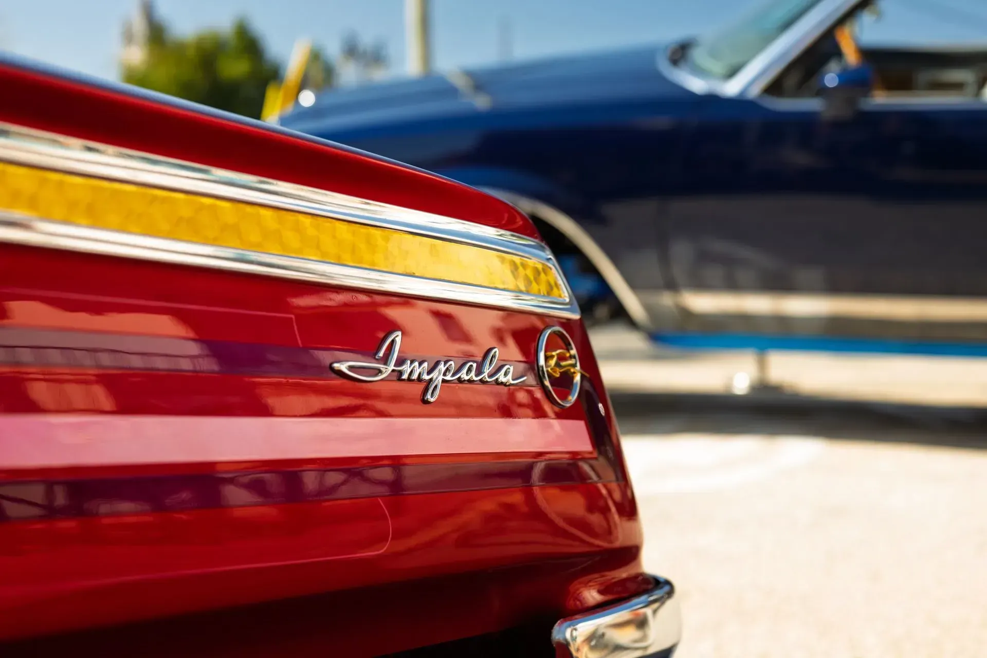 Red car trunk with chrome trim and script badge, parked beside a blue car in sunlight