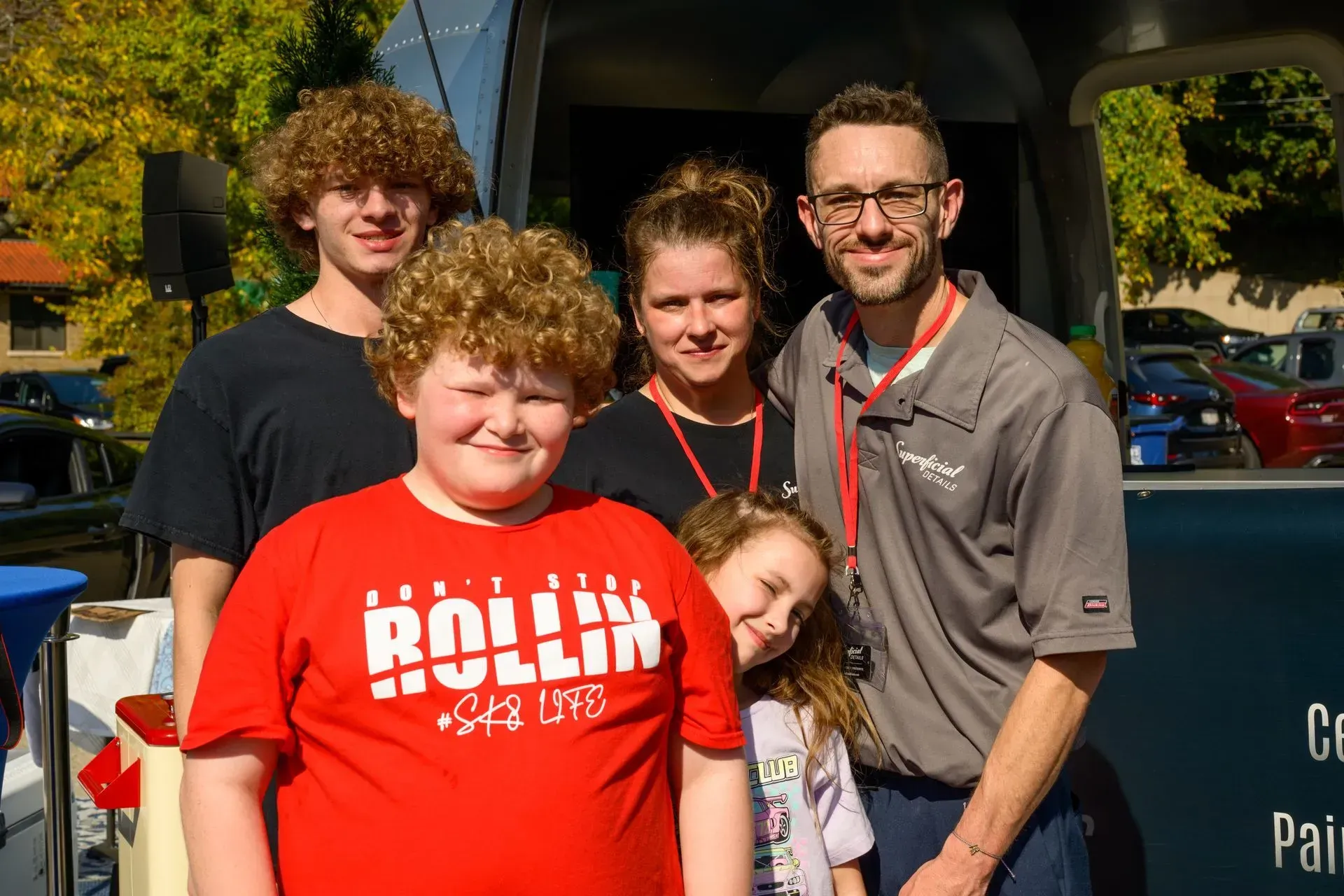 Group of five people posing outdoors in bright sunlight, one in a red “Rolling” shirt and one holding a toddler.