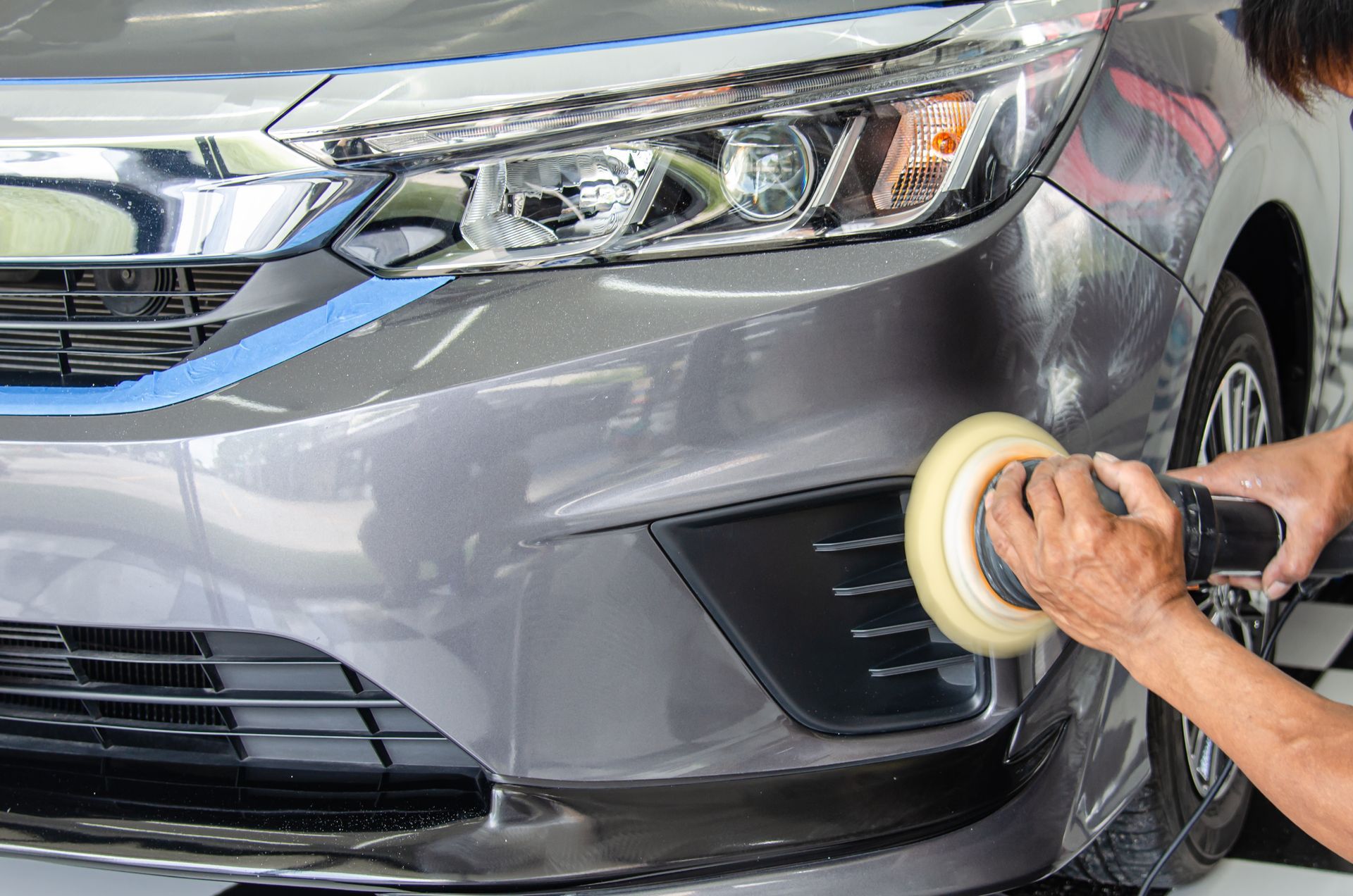 Hands polishing the front bumper of a gray car with a buffing pad