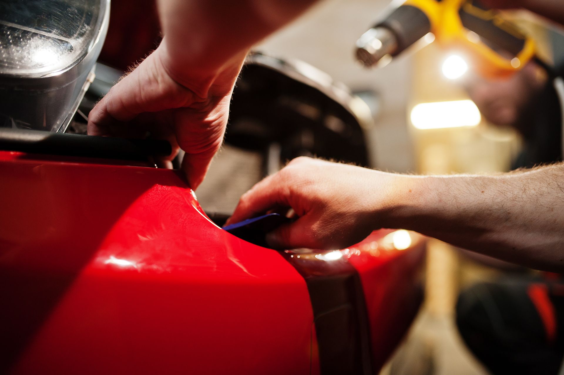 Hands working on a red motorcycle tank in a garage, with tools and blurred background lights