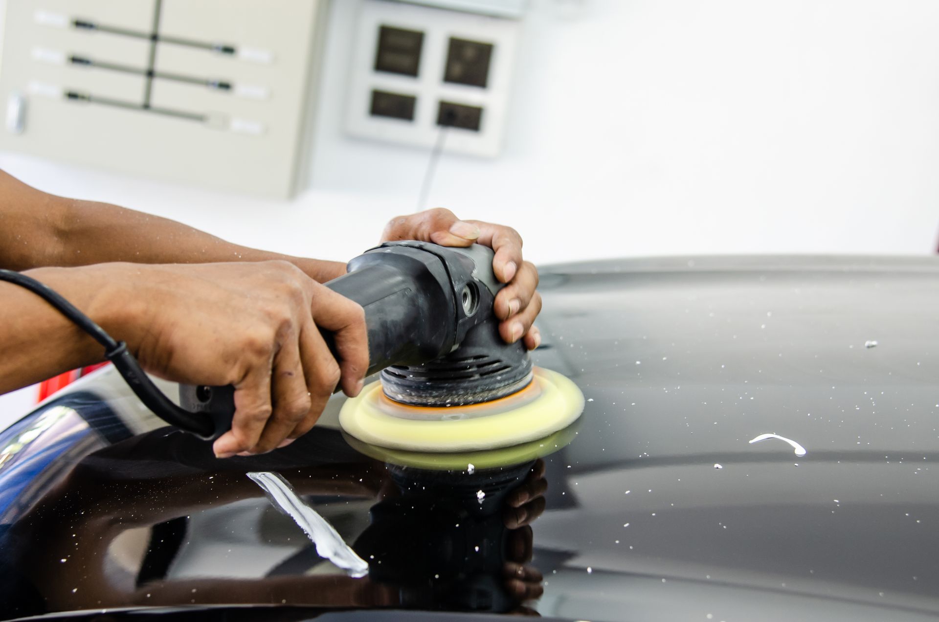 Hands polishing a black car hood with a buffer and yellow pad