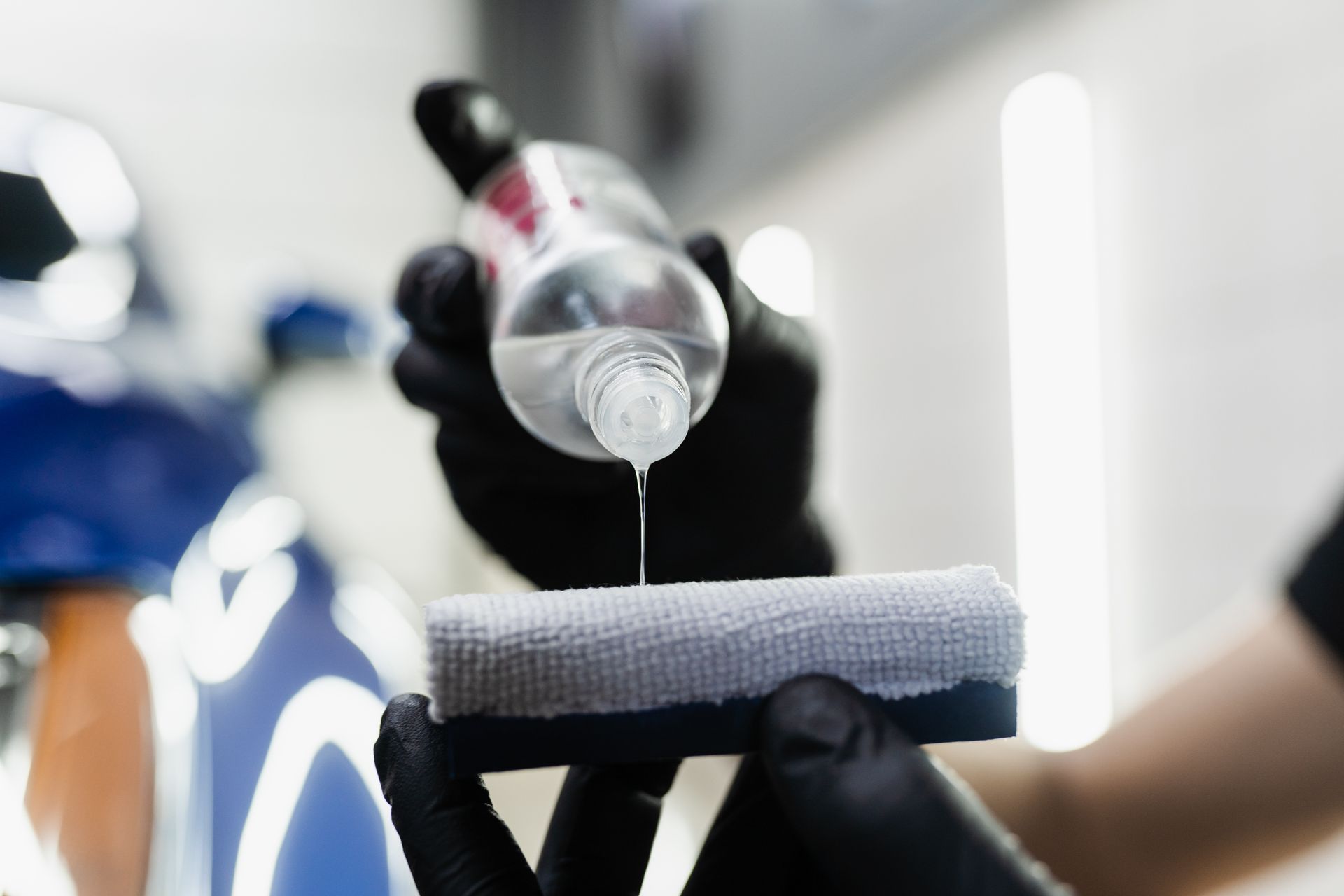 Gloved hands pour liquid from a bottle onto a polishing pad on a car surface