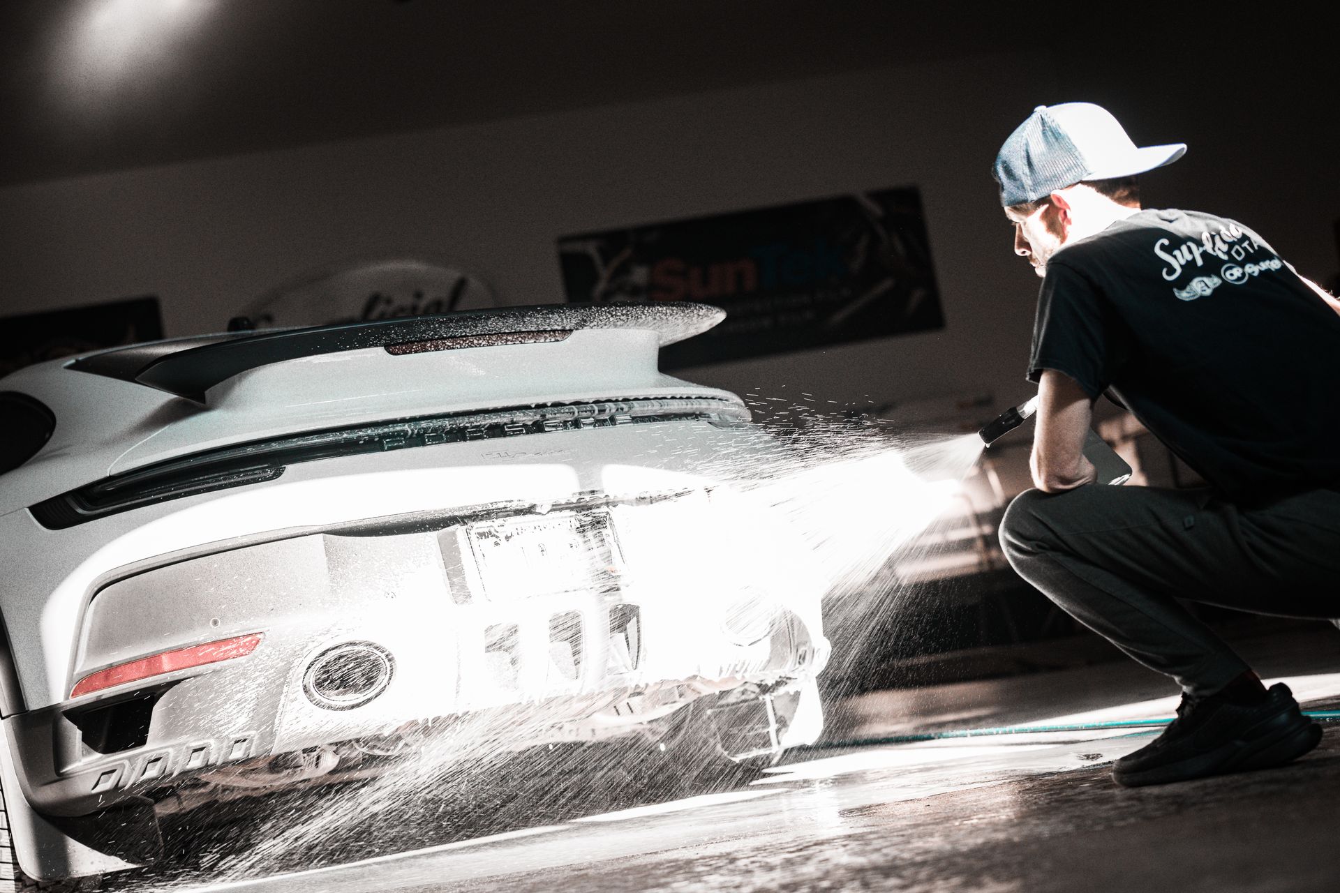 Person pressure-washing a white car in a garage, with water spraying across the rear and side