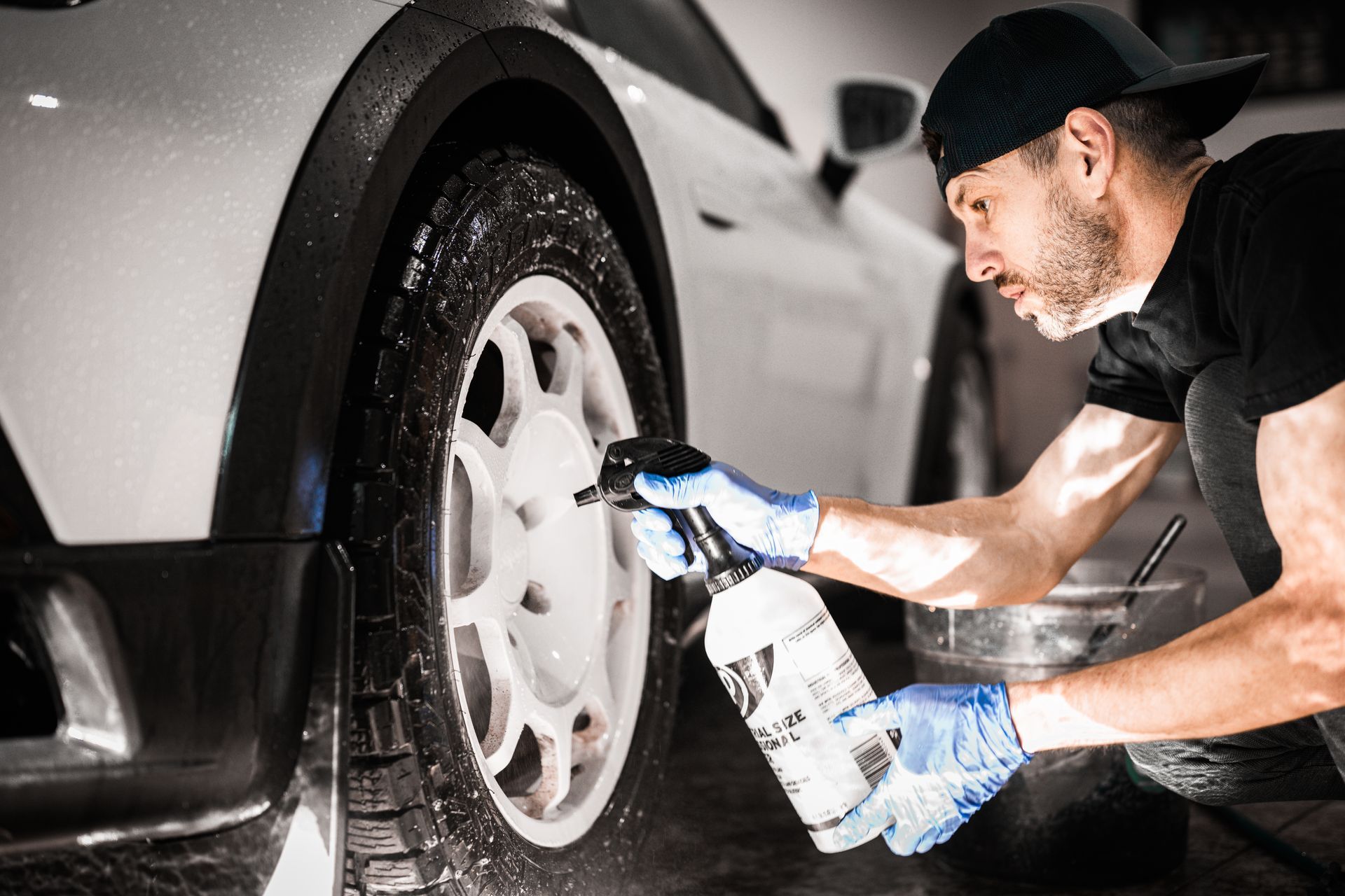 Man washing a silver car wheel with a spray bottle and cloth in bright sunlight
