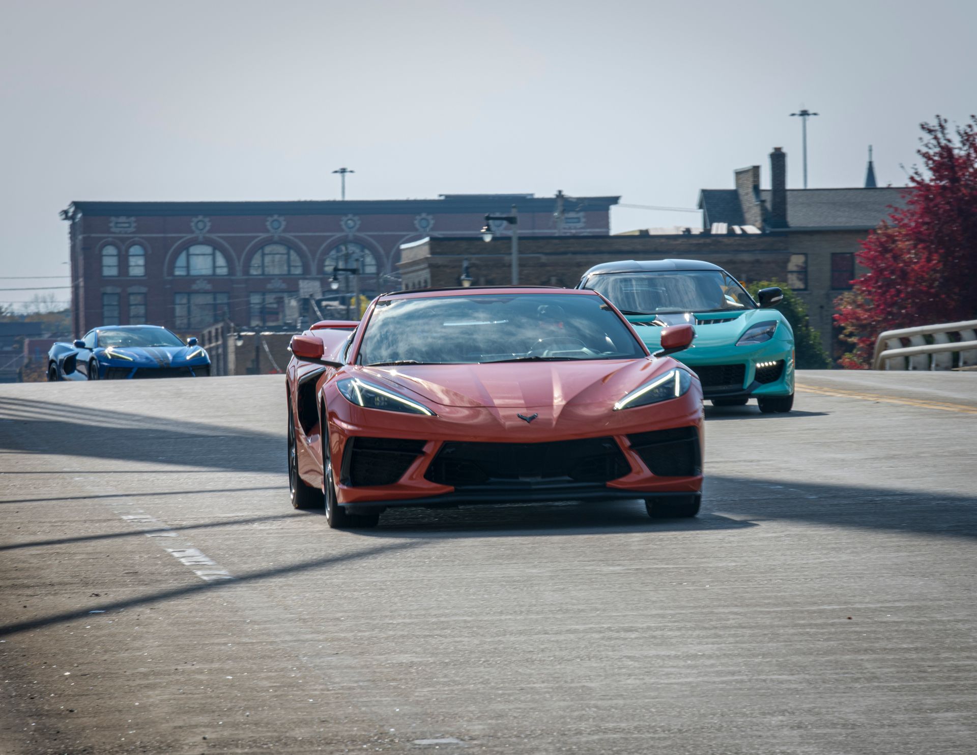 Red sports car leading two cars on a road near a building, with another car in the distance.