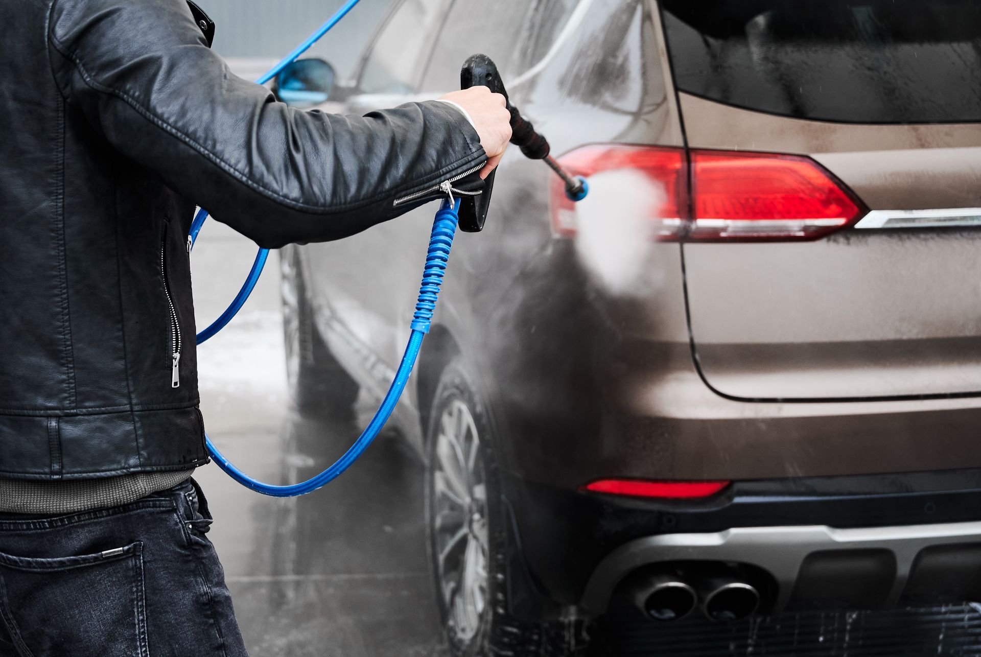 Person pressure-washing the rear of a silver SUV with a blue hose in an outdoor car wash