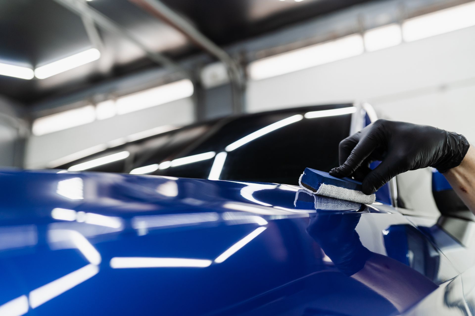 Gloved hand polishing a glossy blue car in a bright auto shop