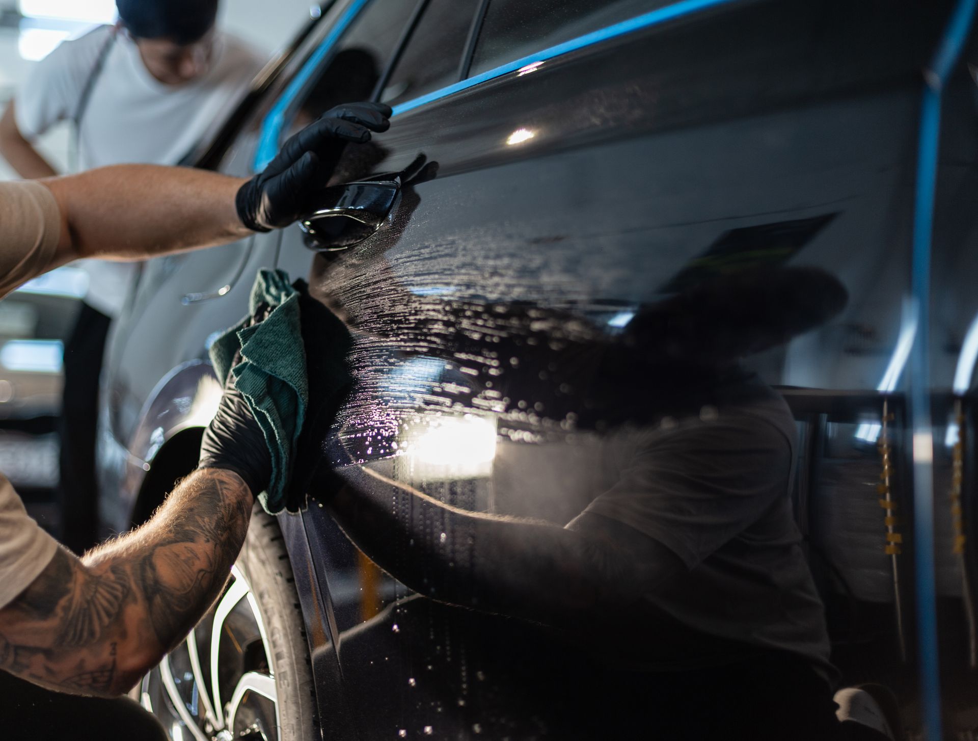 Person cleaning a black car door with a sponge, water spraying in a car wash