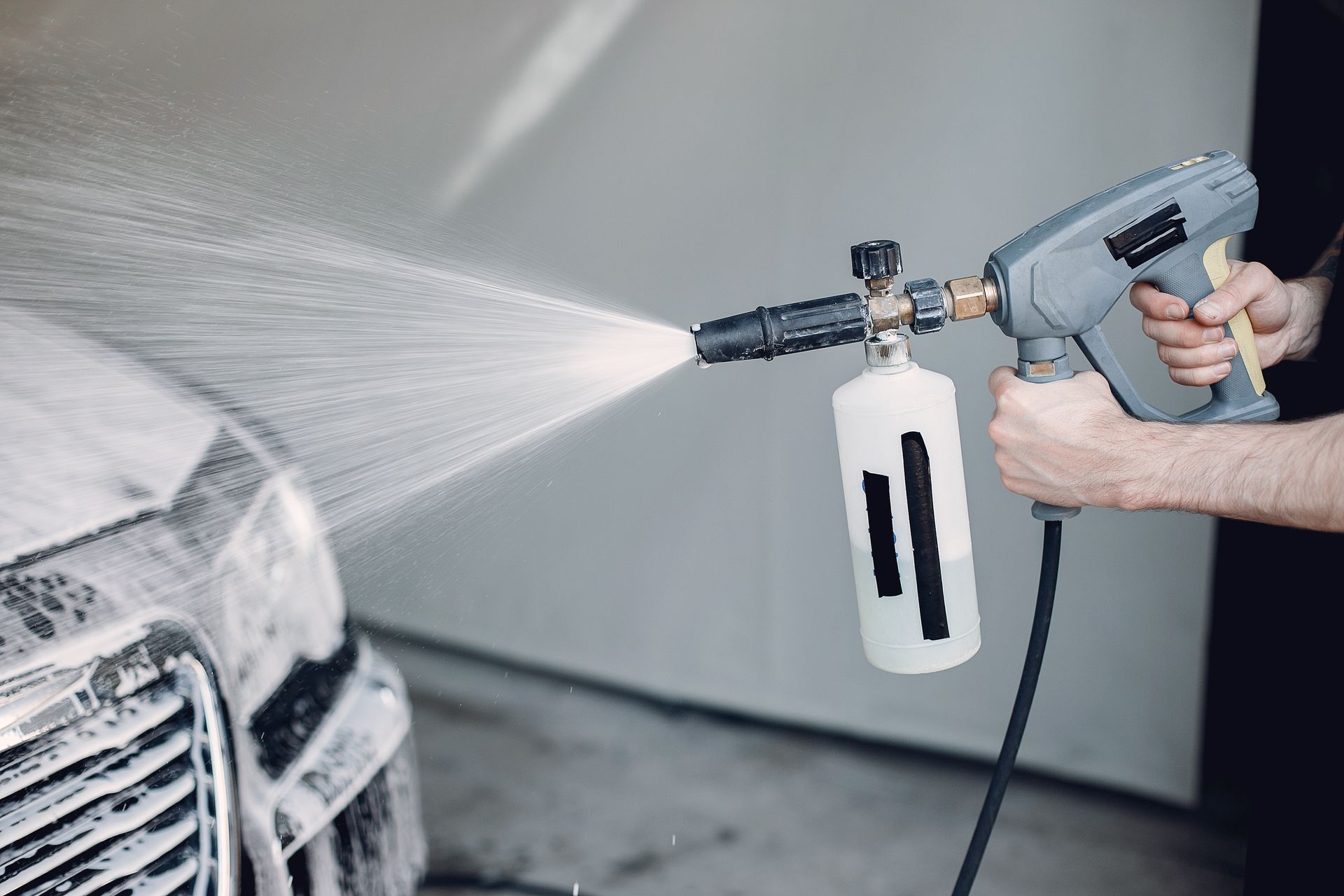 Pressure washer spraying a car’s front end in a garage, with soap bottle attached to the wand