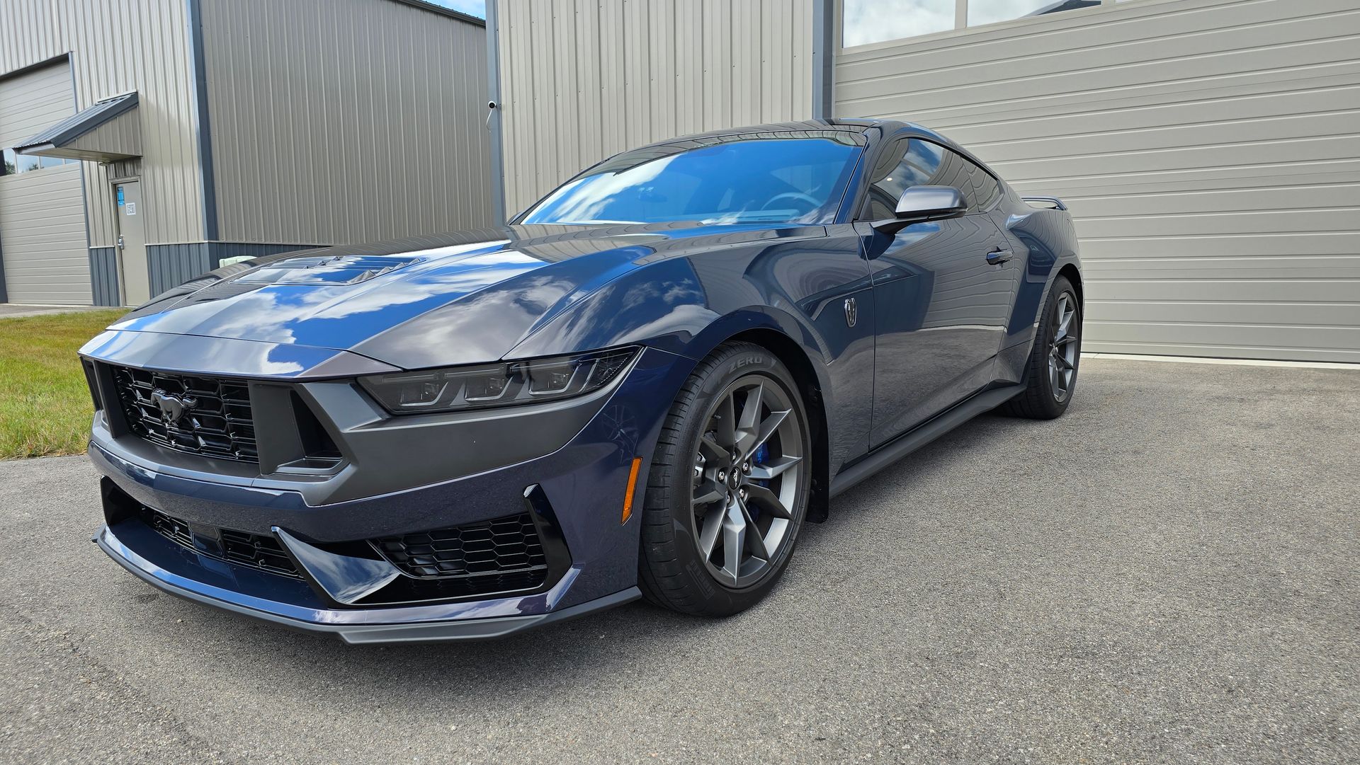 Dark blue sports car parked outside a metal building on a paved lot.