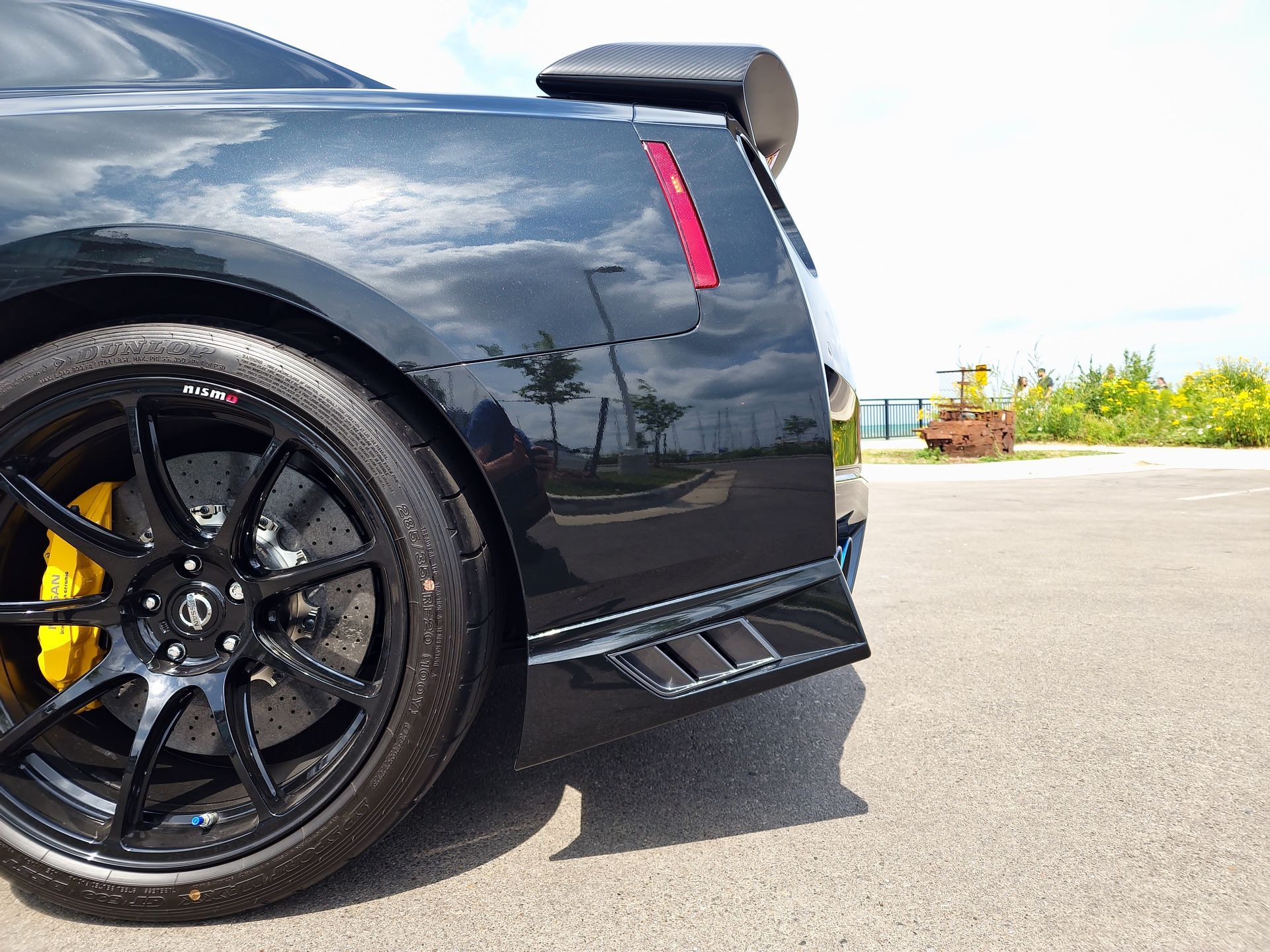 Black sports car rear quarter with glossy paint, black rims, and yellow brake calipers on a sunlit road