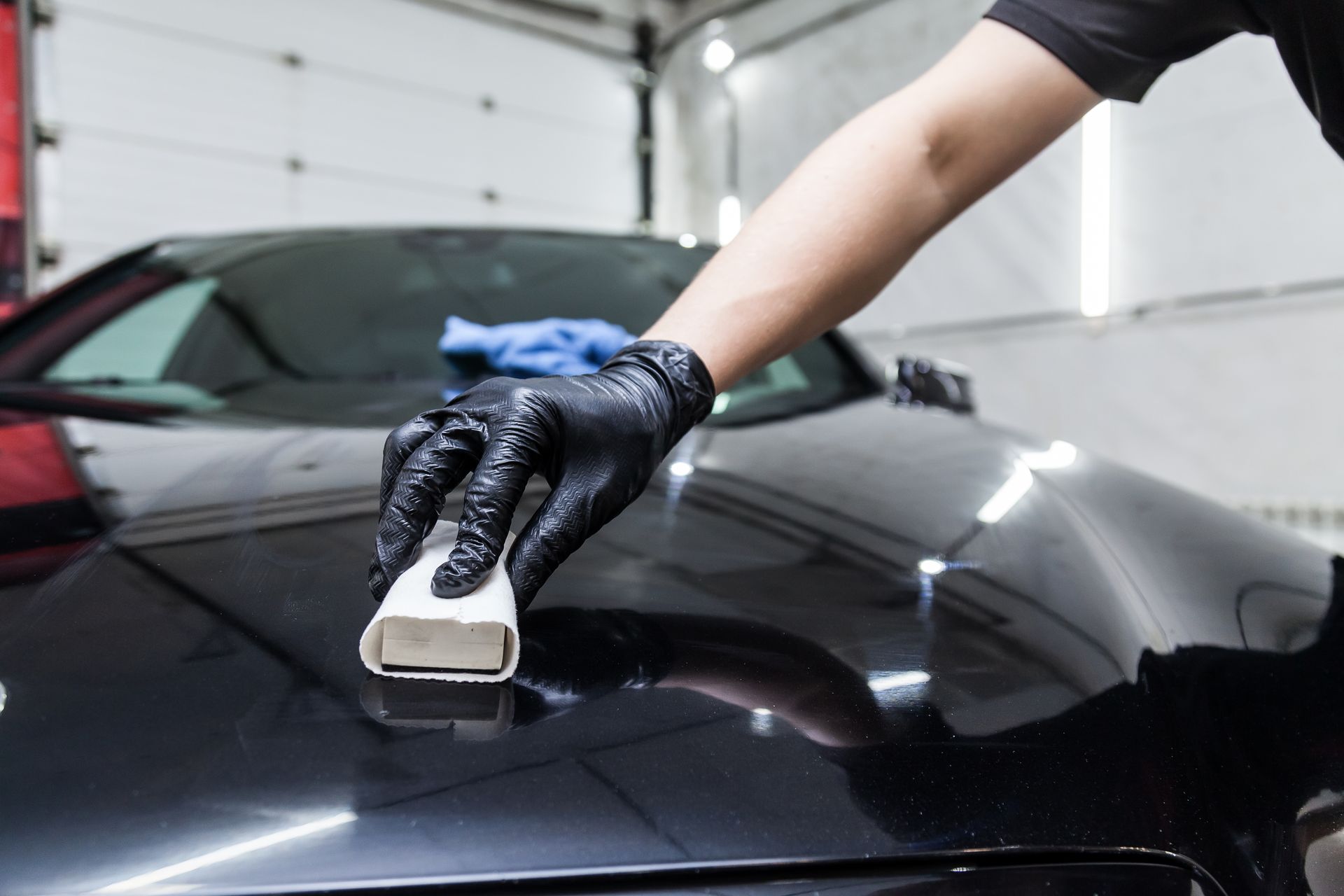 Gloved hand polishing a black car hood with a sponge in a garage