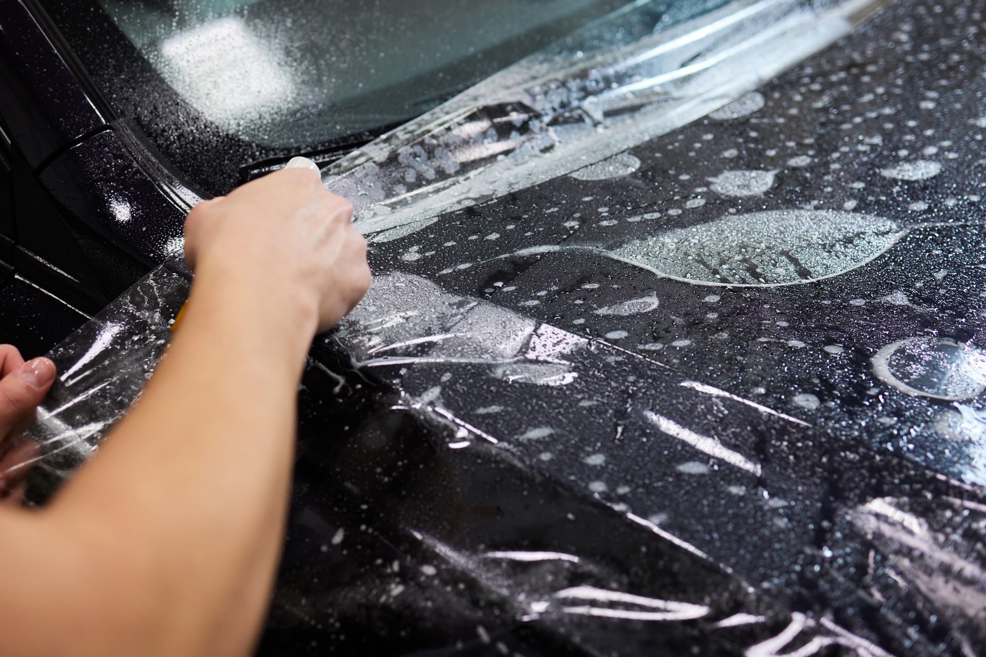 Hands scrubbing soap-covered windshield of a black car with a sponge