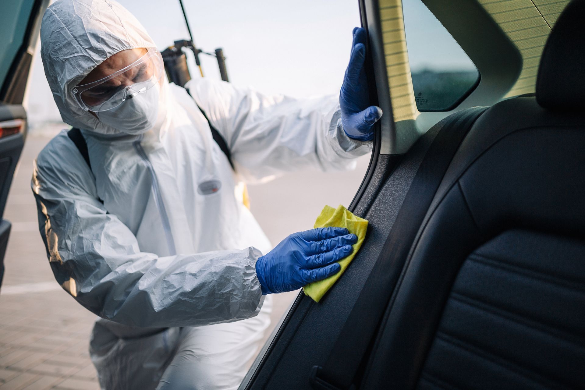 Person in white protective suit cleaning a car door interior with a yellow cloth and blue gloves