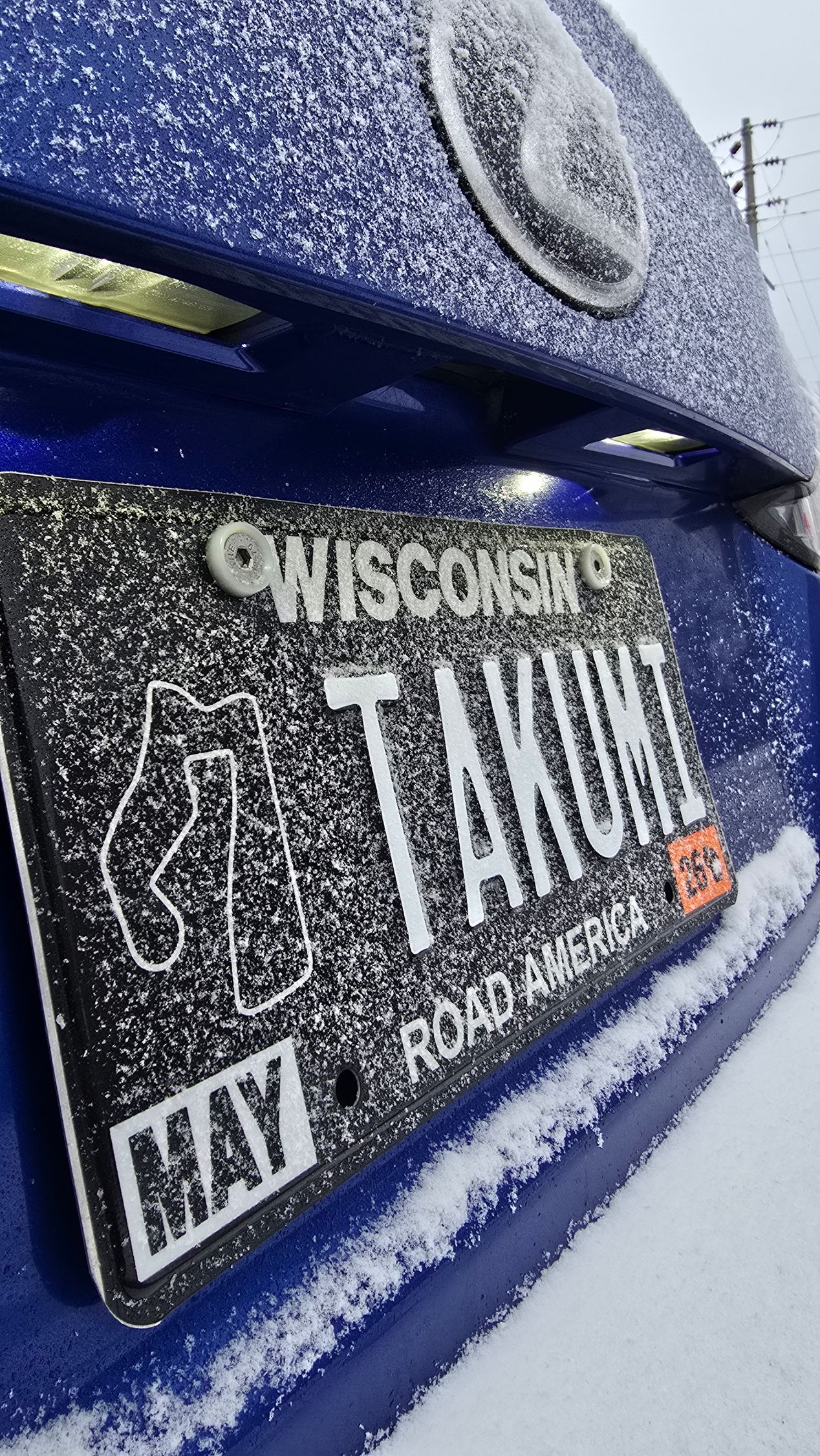 Snow-covered Wisconsin license plate on a blue vehicle, reading “TANU.”