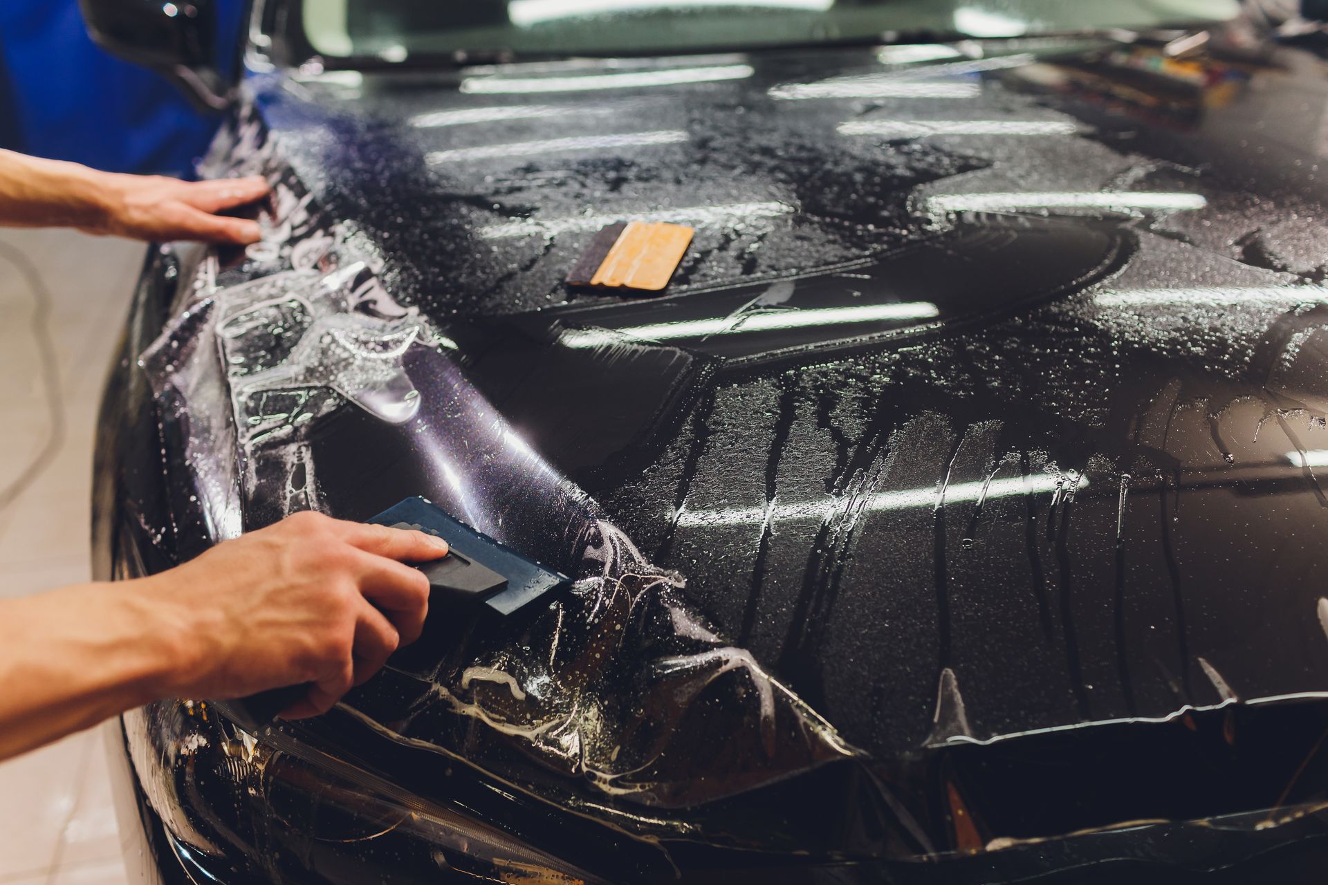 Hands applying clear protective film to a black car hood during detailing